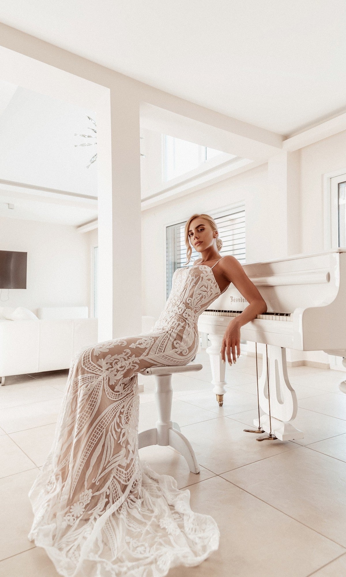 Tina Holly wears the White Lace Bodycon Bridal Gown: BA999, sitting on a white bench beside a white grand piano in a bright, modern room with large windows.